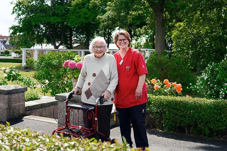 En äldre med rollator och en personal står i en blommig grönska och ler.