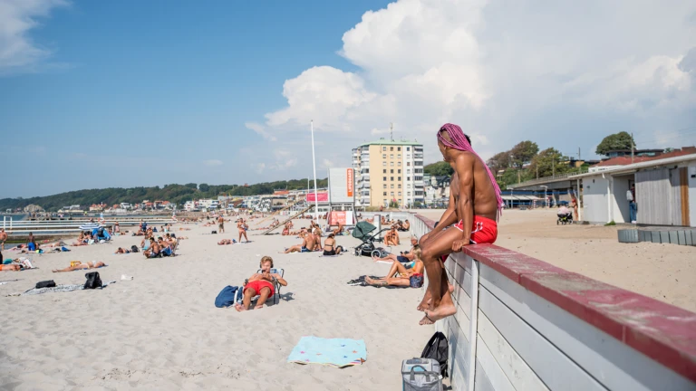 Två personer på Fria bads palissad tittar ut över solbadare på stranden.