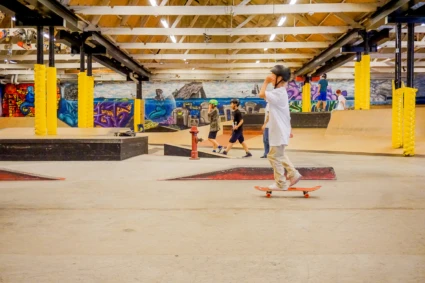 En grupp ungdomar åker skateboard inomhus på en skatepark med färgglada väggmålningar och gulklädda pelare. En person i förgrunden står på en skateboard med hjälm på huvudet. Andra deltagare syns i bakgrunden medan de går eller åker skateboard.