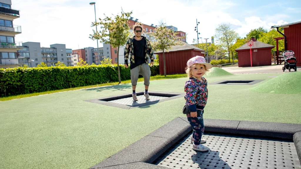 Pappa och dotter hoppar studsmatta på lekplatsen på Furutorpsplatsen.