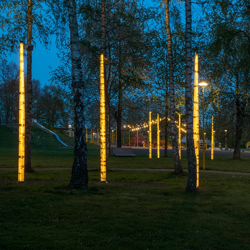 Belysning på björkstammar i Gubbaparken i Helsingborg med en belyst lekplats i bakgrunden.