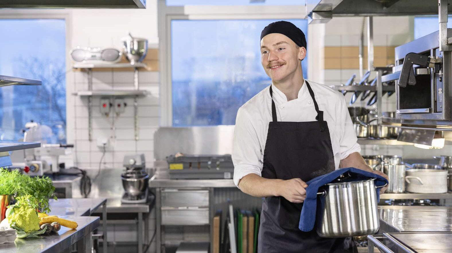 En kock står i ett restaurangkök och bär en kastrull.