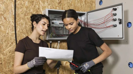 Två elektriker står framför ett öppet elskåp. De pratar och tittar på en ritning som de håller upp framför sig.