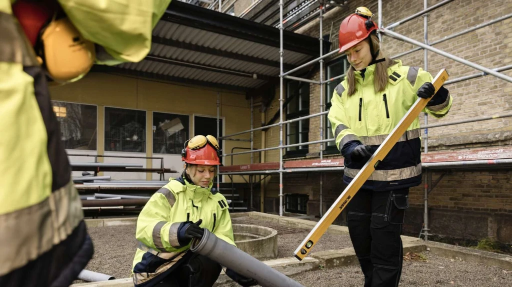 Två vvs-arbetare iklädda varselkläder lägger ner rör i sand.