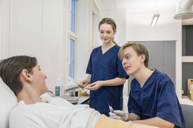 två personer i vårdkläder tar blodprov på patient som ligger i säng.