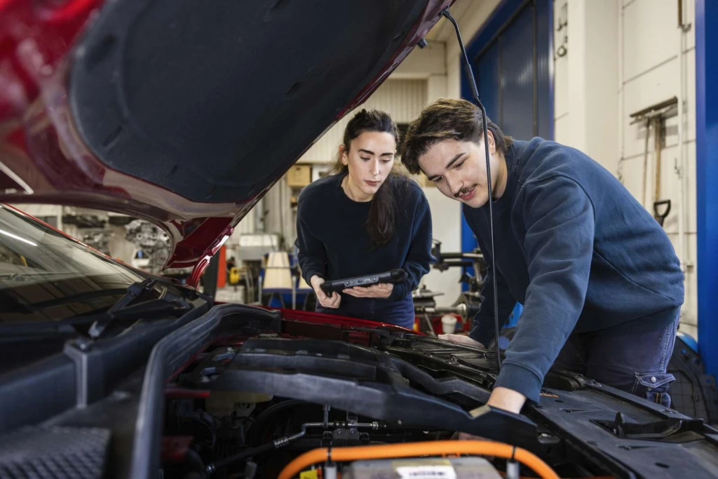 Två personer står vid en öppen motorhuv. En person håller i en surfplatta medan den andra pekar på något i inuti motorn.