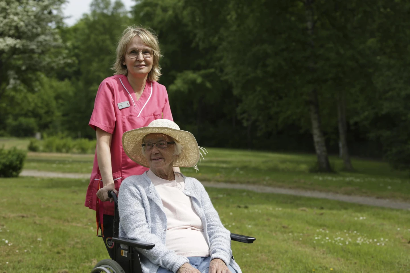 En kvinna sitter i rullstol. Bakom rullstolen står en kvinnlig vårdpersonal.