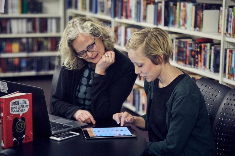 Två kvinnor tittar på en surfplatta på Helsingborgs stadsbibliotek. I bakgrunden syns hyllor med böcker.