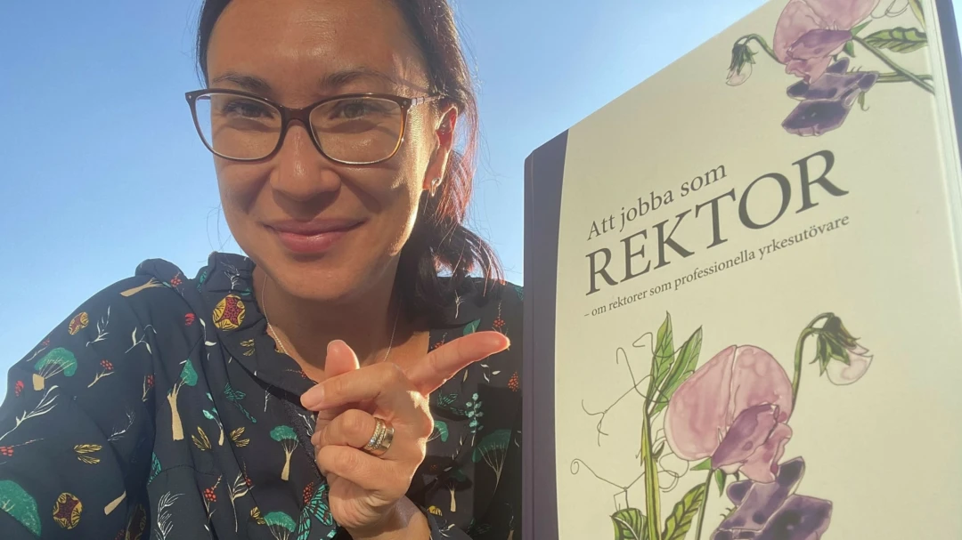 Kvinna med glasögon pekar på boken "Att jobba som rektor: om rektorer som professionella yrkesutövare"