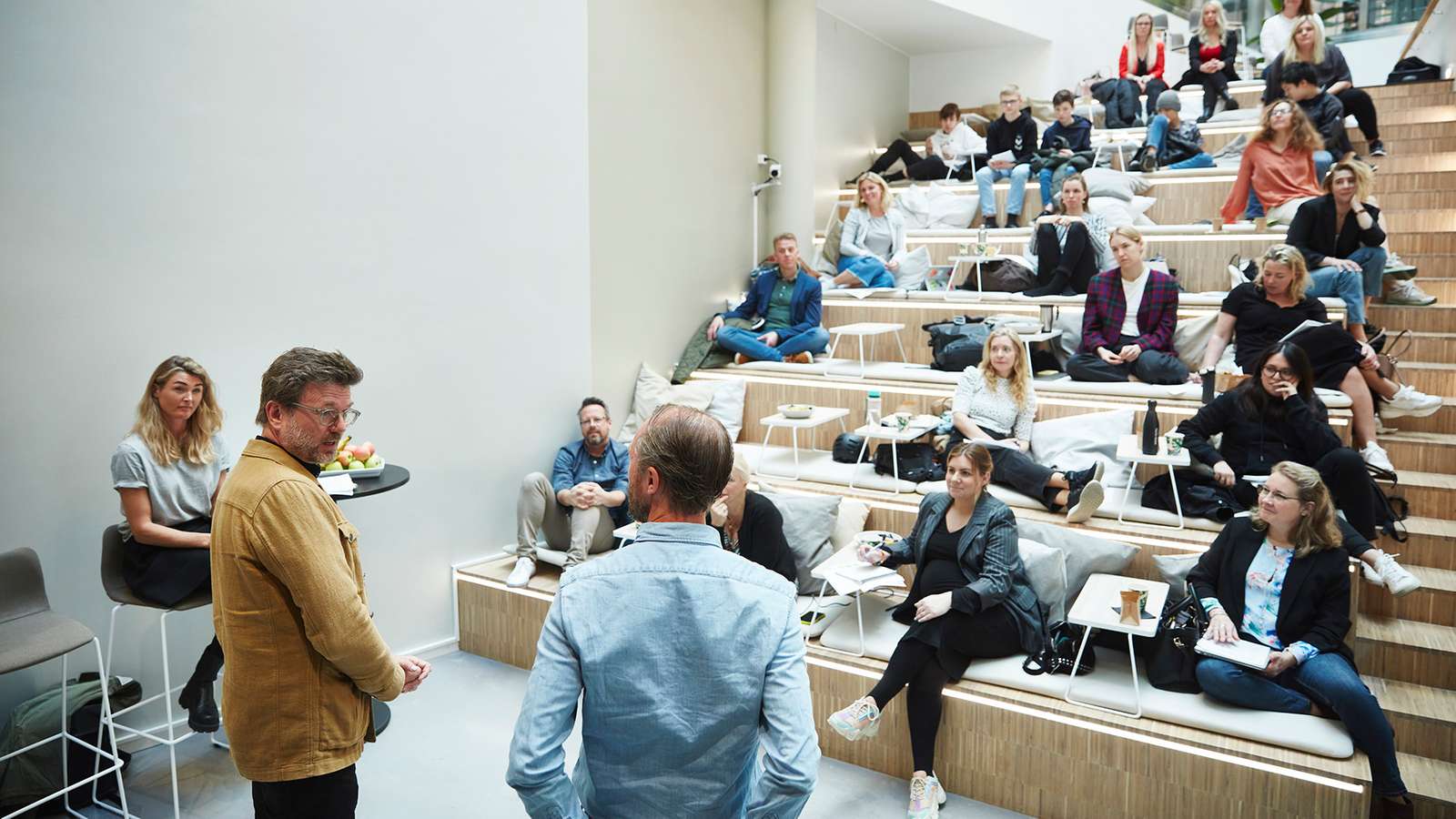 Två personer står och pratar framför en publik, utspridd i en trappliknande läktare.