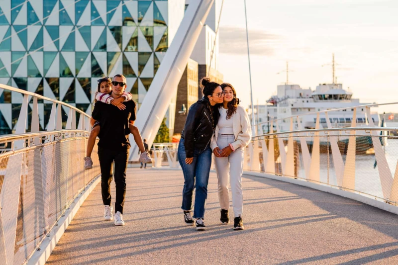 Två vuxna och två barn promenerar över Varvsbron, med Prismahuset och en färja i bakgrunden.