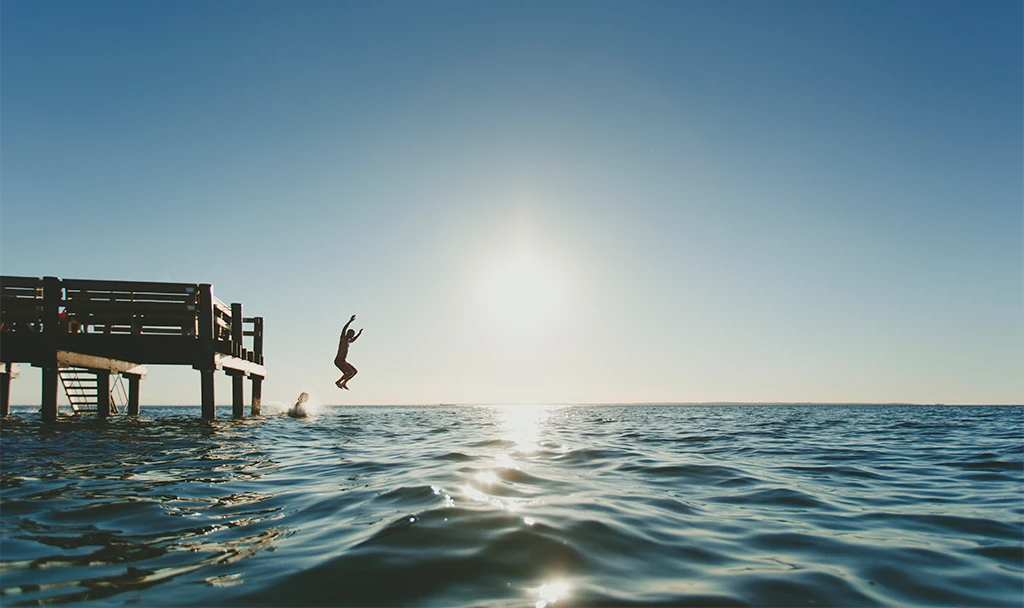 En person hoppar i havet från en brygga, med solen som glittrar i havsytan.