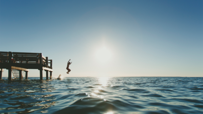 En person hoppar i havet från en brygga, med solen som glittrar i havsytan.
