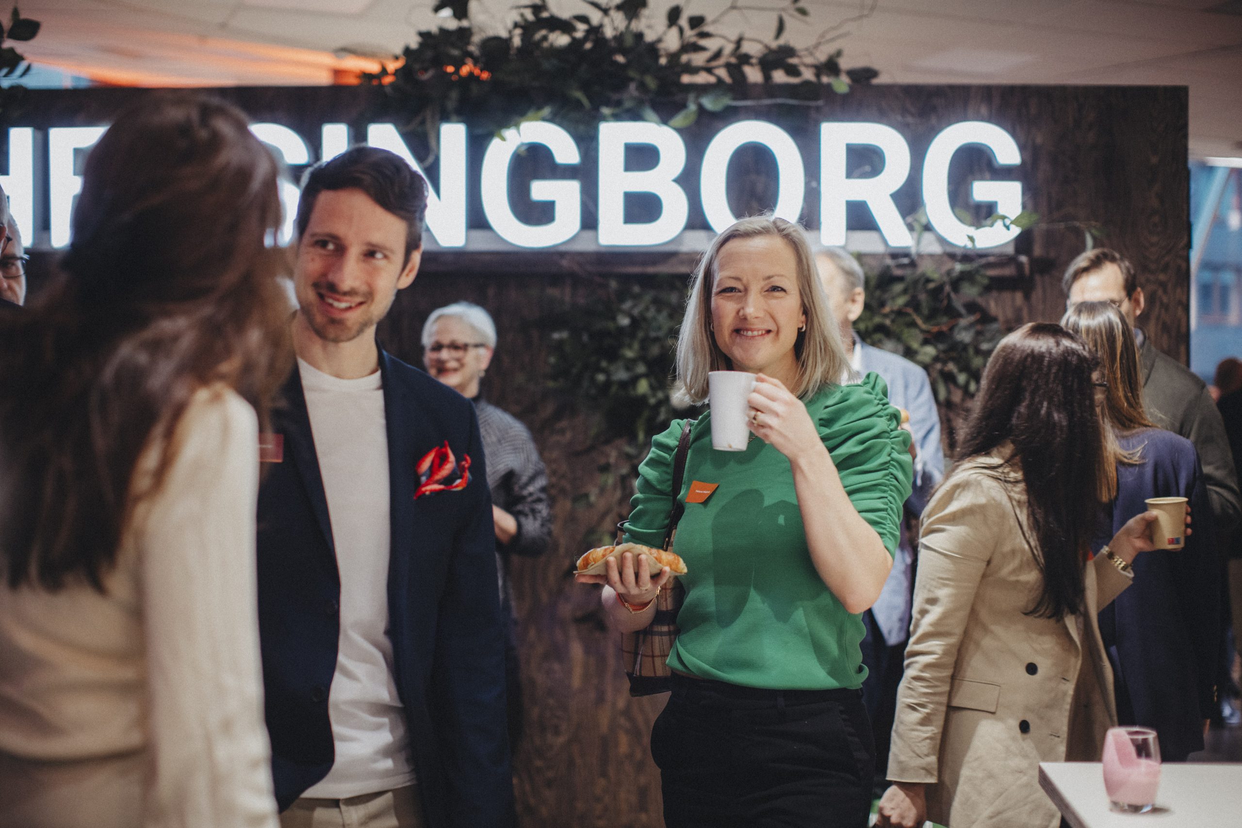 Två personer står i ett minglande folkhav, den ena med kopp och bakverk i handen. I bakgrunden en stor text som säger "Helsingborg".