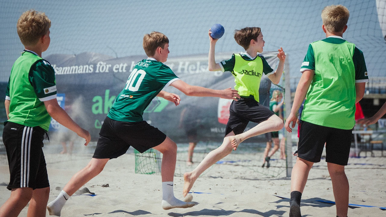 Fyra killar sim spelar beachhandboll i gröna tröjor och västar på stranden i Helsingborg.