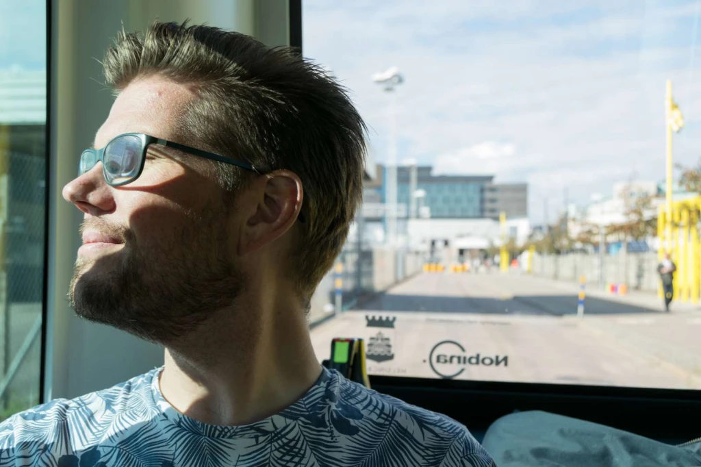 En man med glasögon sitter längst bak i en buss med solen i ögonen.