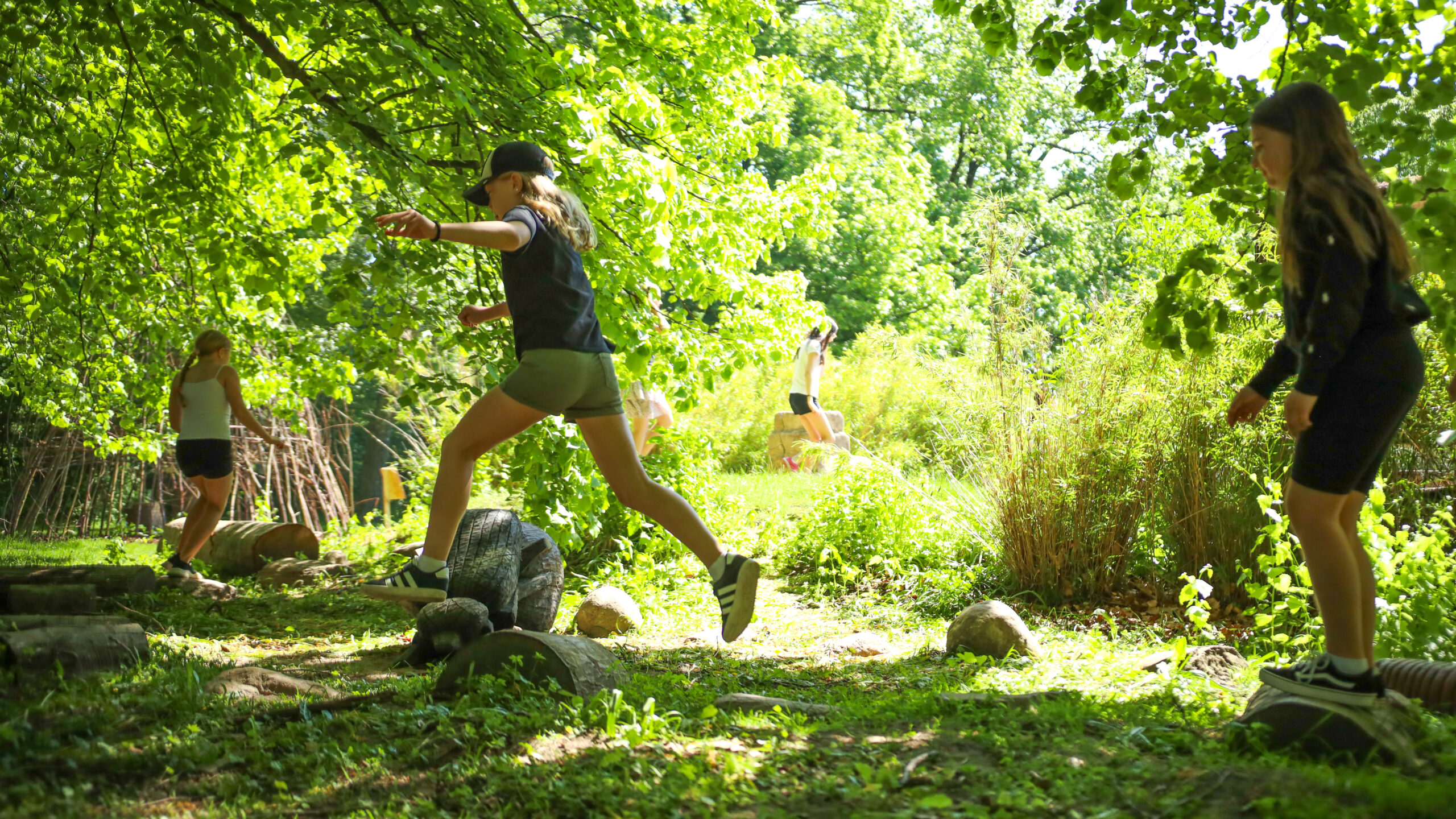 Barn som hoppar mellan hinder på en hinderbana i en solig lövskog.