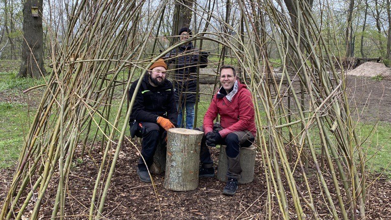 Tre personer som står i en koja av smala grenar från ett pilträd.