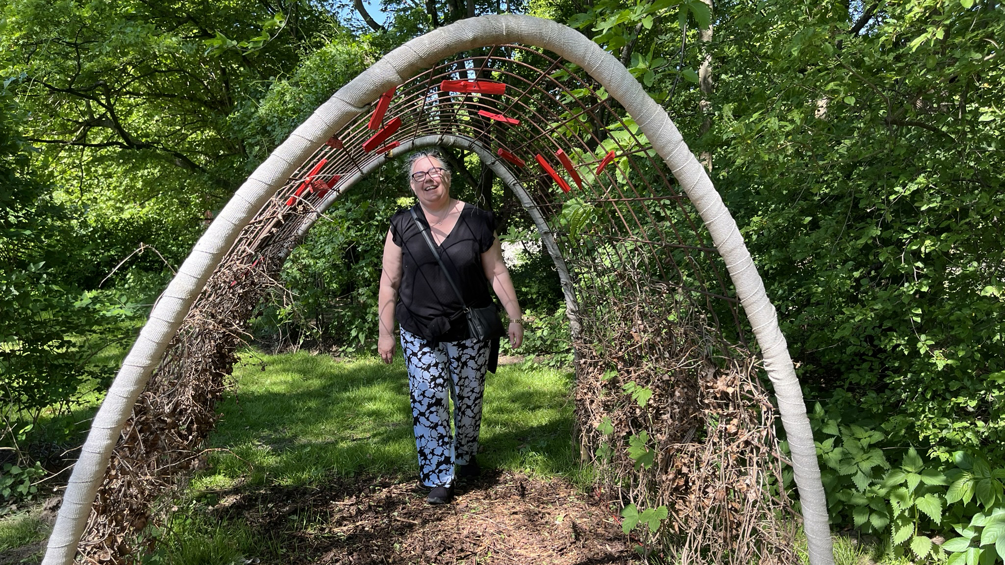 En kvinna går genom en tunnel av ett böjt armeringsnät, där humleplantor slingar sig uppåt längs sidorna.