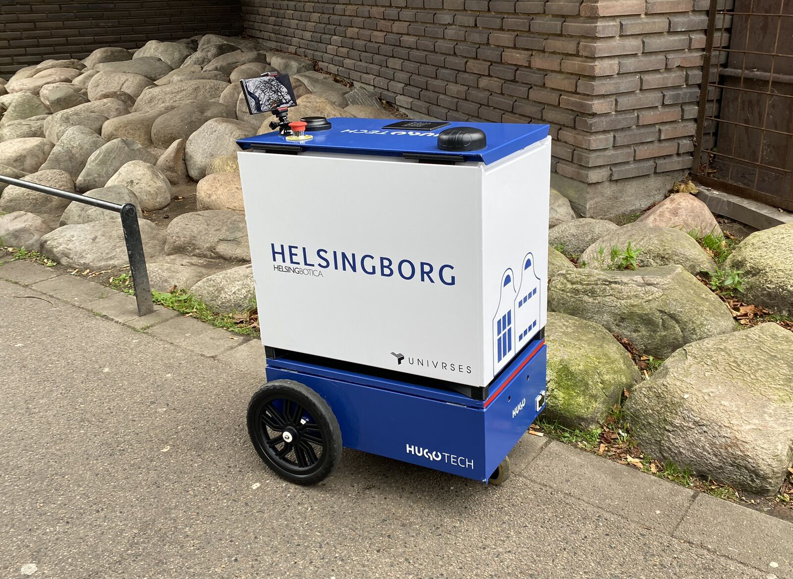 En lådliknande robot på hjul, med en mobiltelefon och en stor, röd stoppknapp på toppen.