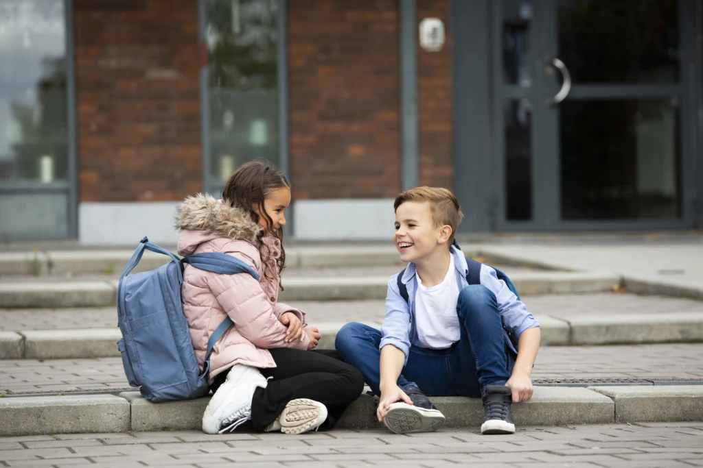 Två barn sitter på trappsteg utomhus framför en byggnad med glasdörrar och tegelvägg.