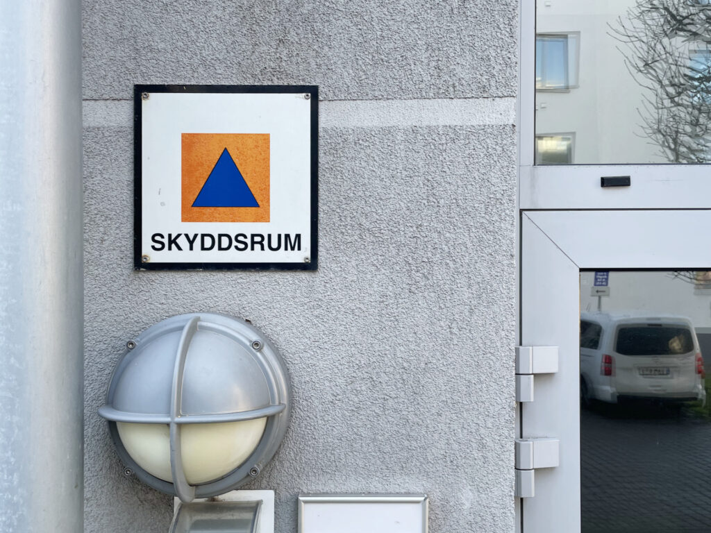 En skylt för skyddsrum sitter på en grå fasad intill en entrédörr