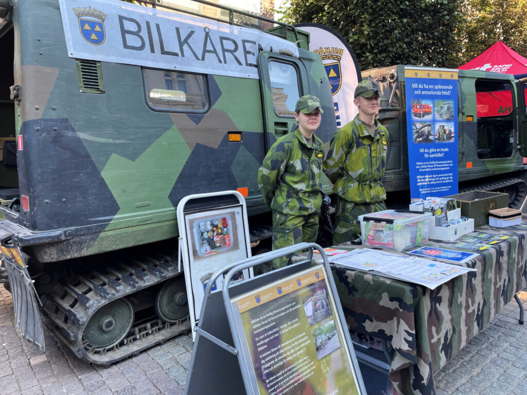 Två personer i grön kamouflageuniform står framför en bandvagn med texten Bilkåren. Framför dem ett bokbord.