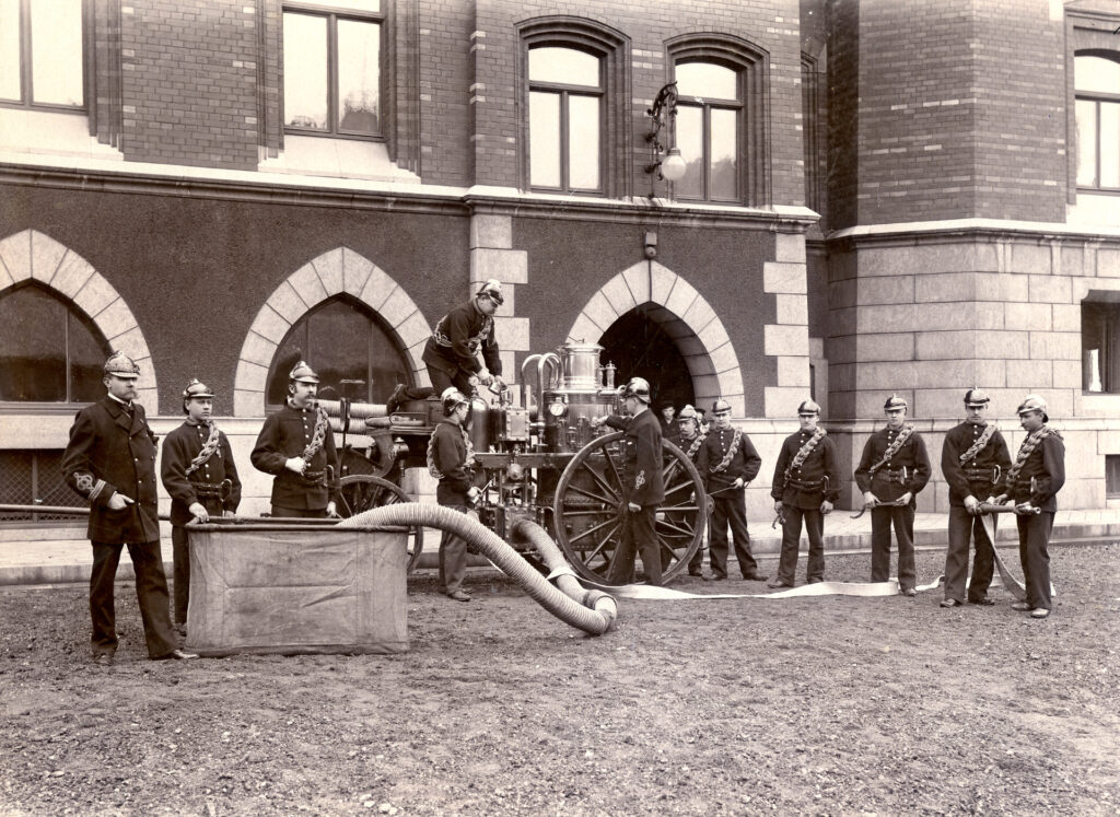 Brandmän står framför Rådhuset med gammal släckningsutrustning och slang. Svartvit bild från 1905.