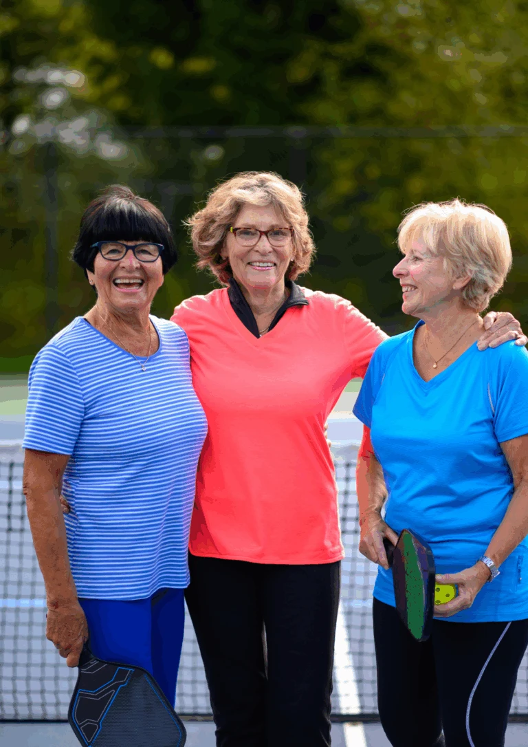 Tre glada kvinnliga seniorer står på en tennisplan och håller om varandra.