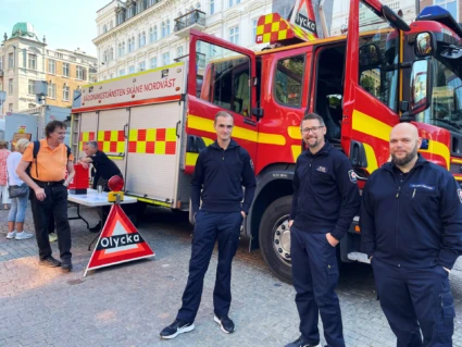 Tre brandmän står framför en släckbil med öppna dörrar på ett torg