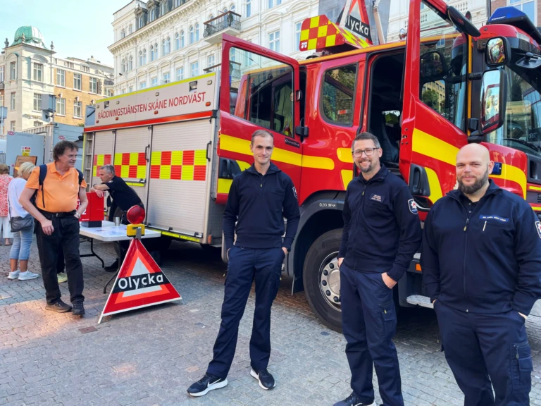 Tre brandmän står framför en släckbil med öppna dörrar på ett torg