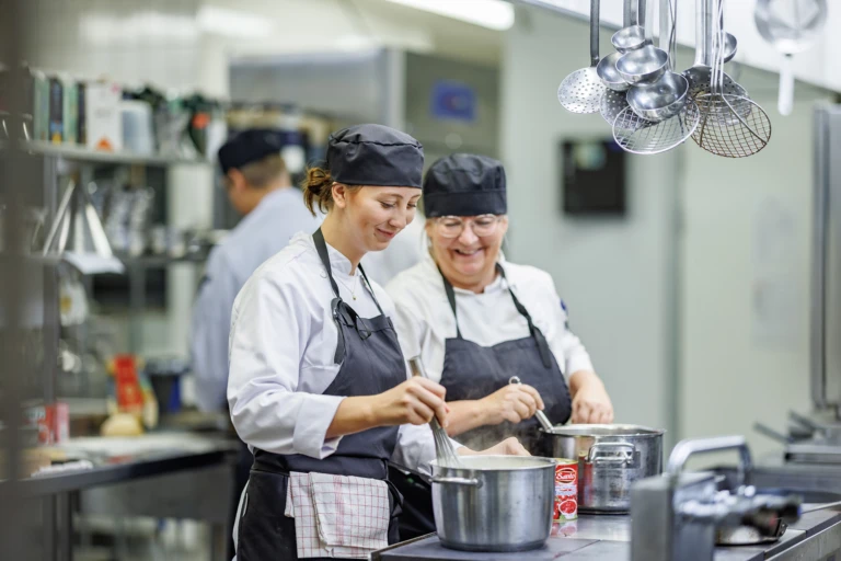 En ung och en äldre person, iklädda kockkläder lagar mat tillsammans i ett kök. Båda ler