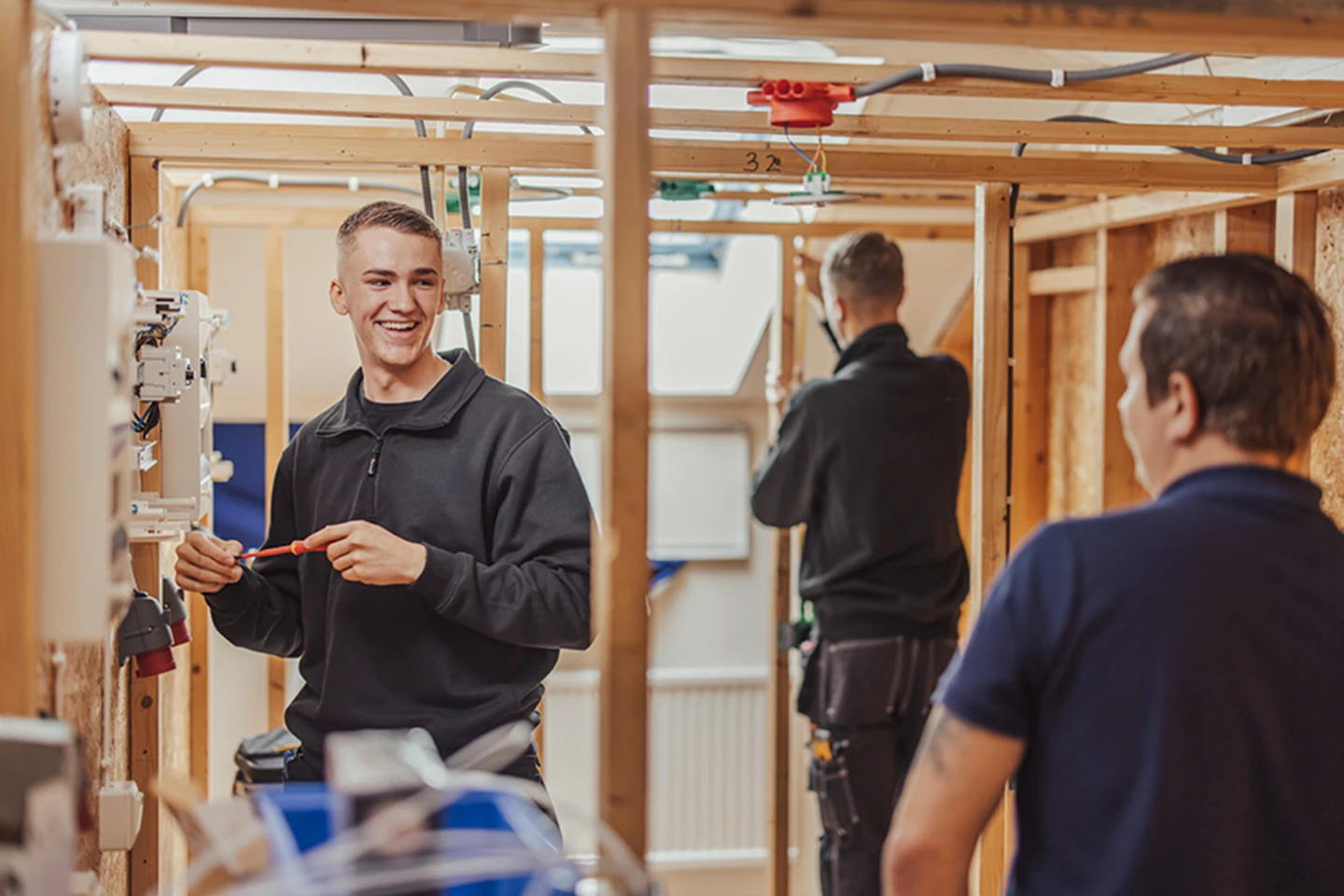 en leende person håller i en skruvmejsel och står bredvid ett elskåp. Personen tittar på en lärare om syns i förgrunden.