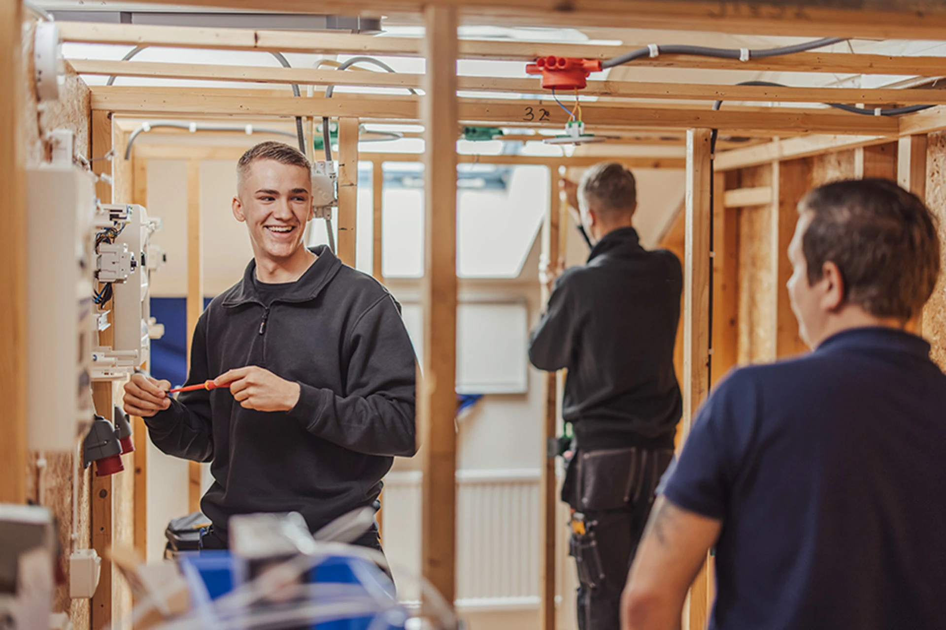 en leende person håller i en skruvmejsel och står bredvid ett elskåp. Personen tittar på en lärare om syns i förgrunden.