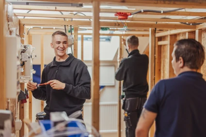 en leende person håller i en skruvmejsel och står bredvid ett elskåp. Personen tittar på en lärare om syns i förgrunden.