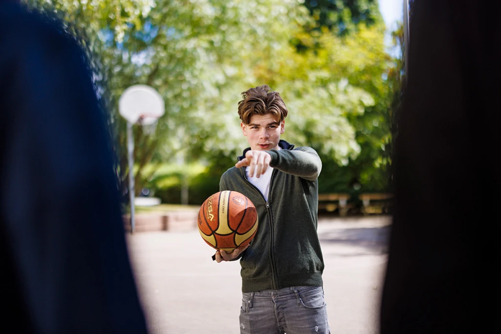 en ung kille håller i en basketboll och pekar in i kameran. I förgrunden synd två personer som står vända mot honom