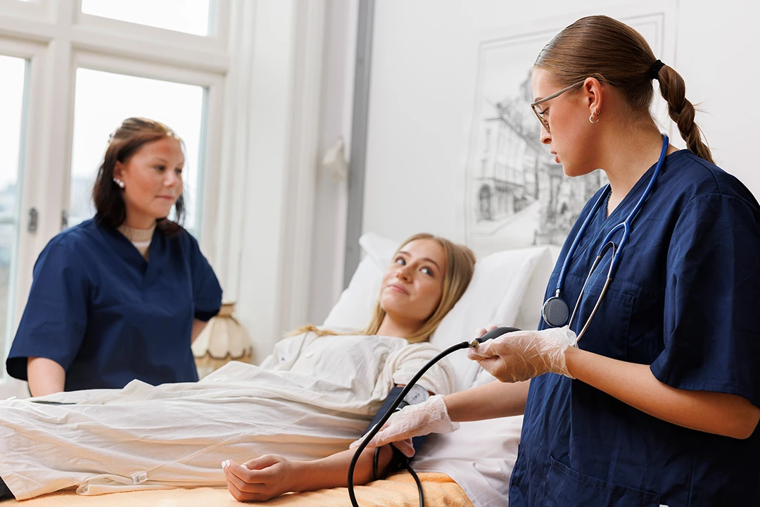 Två person i vårdkläder står bredvid en patient som ligger i en sjukhussäng. Den ena sköterskan tar blodtryck på patienten.