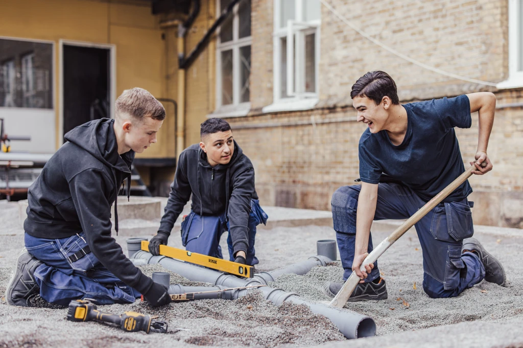 Tre ungdomar iklädda blåställ lägger ner rör i marken samtidigt som de mäter med vattenpass. De ser glada ut.