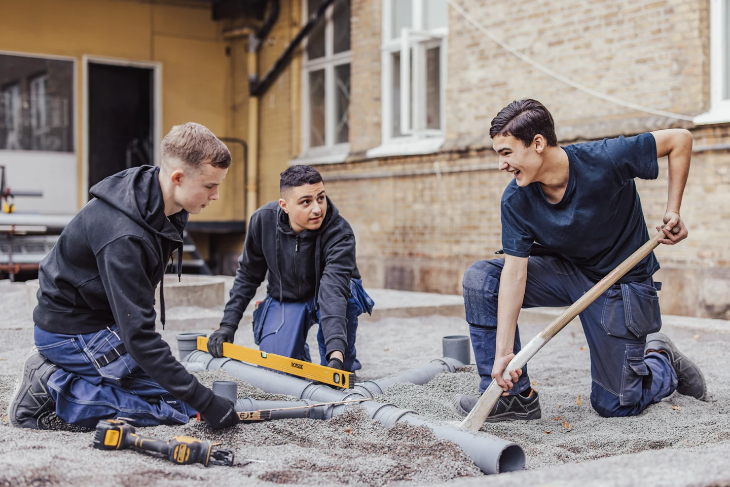 Tre ungdomar iklädda blåställ lägger ner rör i marken samtidigt som de mäter med vattenpass. De ser glada ut.