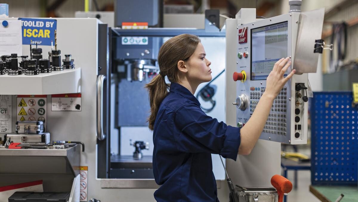 Ung kvinna står framför CNC-maskin. Hon ser koncetrerad ut medan hon trycker på inställningar på datorskärmen.
