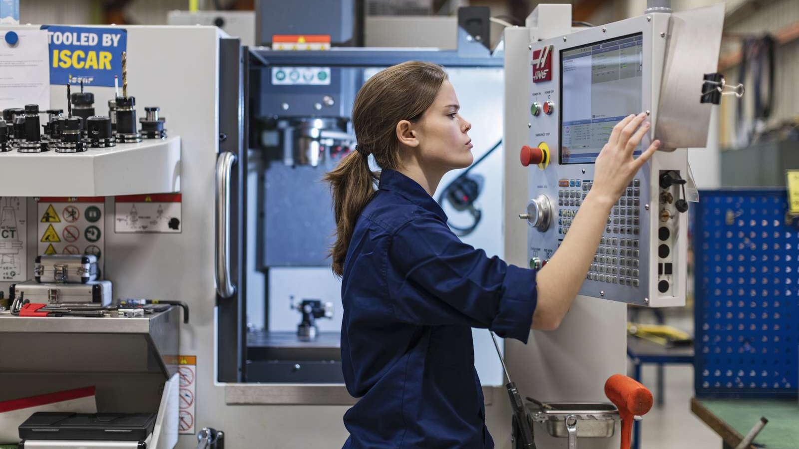 Ung kvinna står framför CNC-maskin. Hon ser koncetrerad ut medan hon trycker på inställningar på datorskärmen.