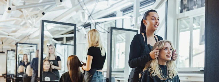 Flera frisörer arbetar med kunder. Frisören närmast fixar kundens hår medan både två ler och tittar in i spegeln framför