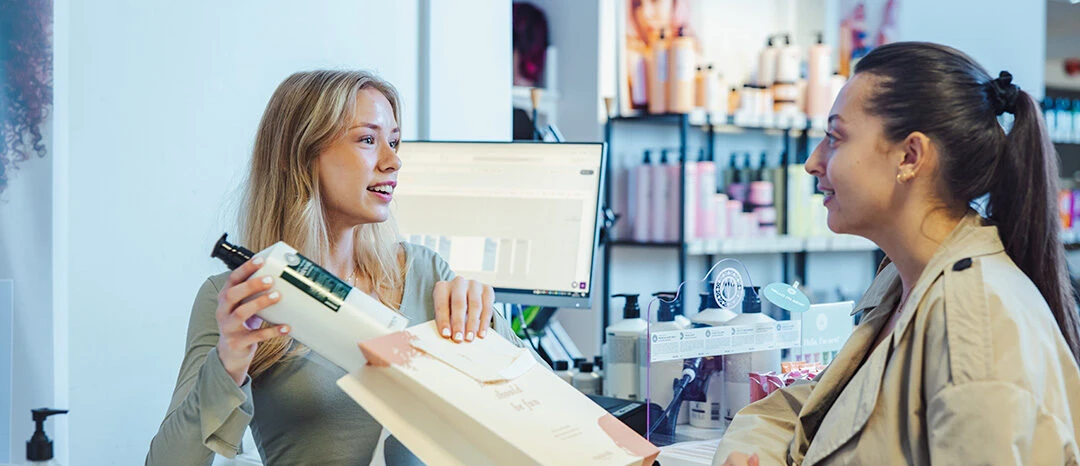 En peson står bakom en kassa och packar ner en flaska i en påse. Samtidigt tittar hon på och ler mot en kund som står framför