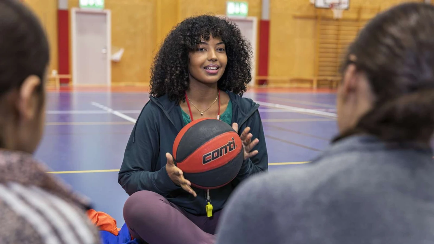 Tränare sitter på golvet i en gympasal och håller i en basketboll. I förgrunden syns två personer i träningskläder.
