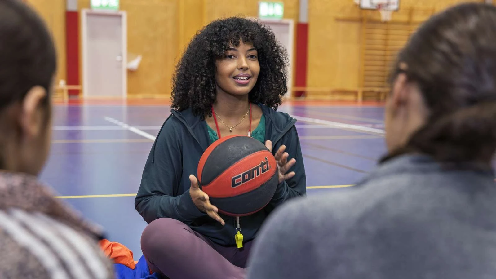 Tränare sitter på golvet i en gympasal och håller i en basketboll. I förgrunden syns två personer i träningskläder.
