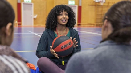 Tränare sitter på golvet i en gympasal och håller i en basketboll. I förgrunden syns två personer i träningskläder.