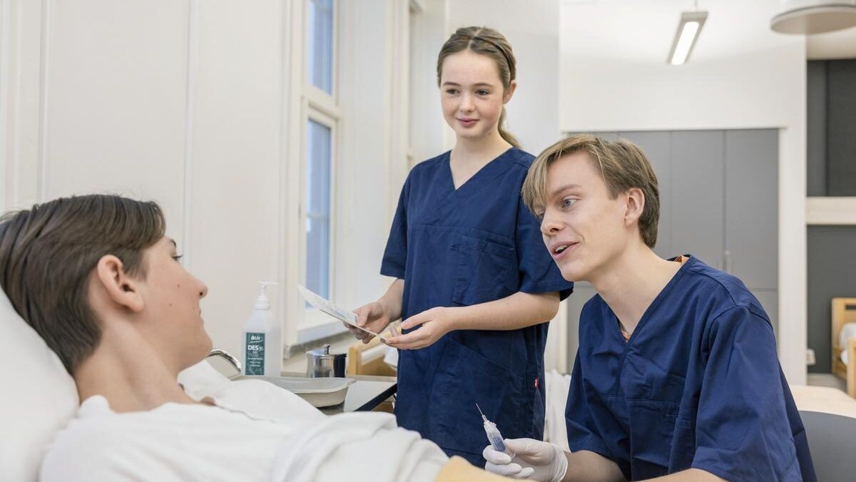 två personer i vårdkläder tar blodprov på patient som ligger i säng.