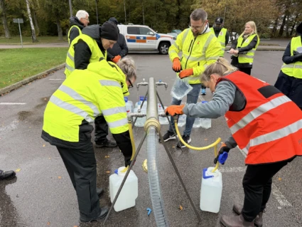 Flera personer i varselvästar tappar vatten i vattendunkar från ett metallrör på en asfaltsyta