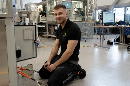 Alvin sitter på knä framför en maskin i en fabrik. Ur maskinen löper elkablar och i handen håller Alvi en tång