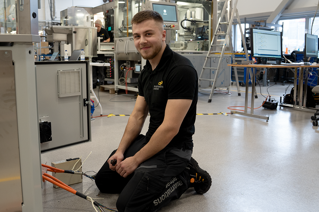 Alvin sitter på knä framför en maskin i en fabrik. Ur maskinen löper elkablar och i handen håller Alvi en tång